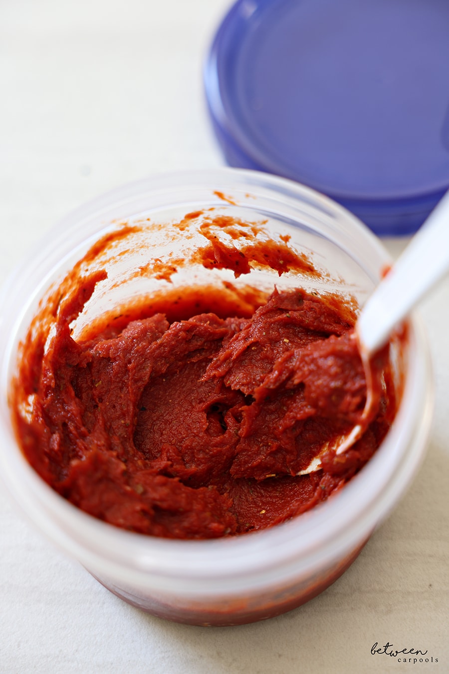 An open 16 oz twist top storage container with tomato paste and a plastic spoon.