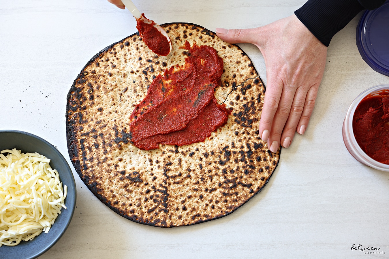 Spreading a dollop of seasoned tomato paste on one round Shmurah Matzah with a plastic spoon. An open 16 oz twist top storage container with seasoned tomato paste and a bowl with shredded cheese nearby.